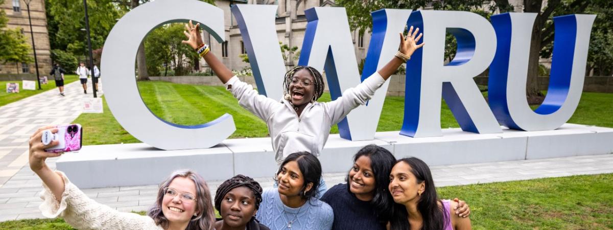 Five students stand in front of the CWRU signage to take a selfie while a sixth jumps with her arms splayed behind them.