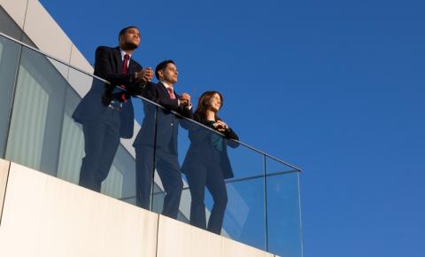 Three individuals in business attire stand on an outdoor balcony looking out.