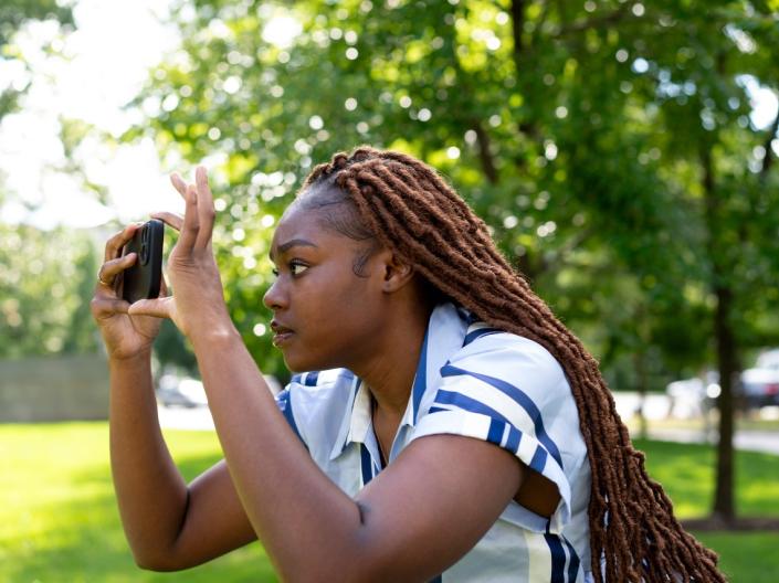 A student focuses intently on her phone screen while taking a picture outdoors.