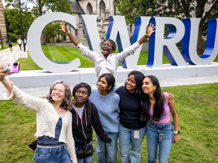 Five students stand in front of the CWRU signage to take a selfie while a sixth jumps with her arms splayed behind them.