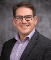 Headshot of Marvin Nieman in a blue collared shirt black jacket and grey blurred background