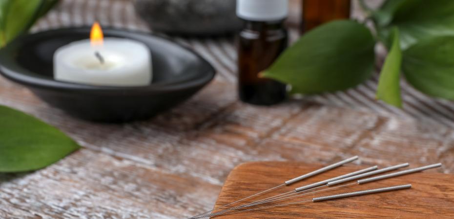 Acupuncture needles on a piece of wood on a table with a lit candle in bowl, leaves, stones and bottles in the background.