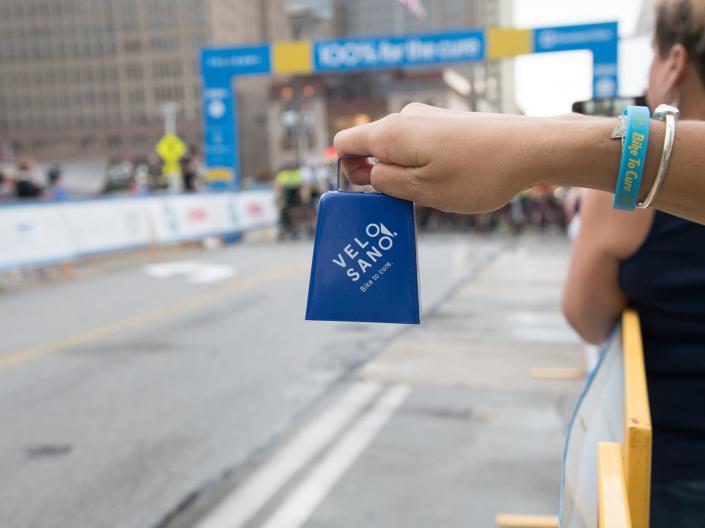 Person holding cowbell with VeloSano bike ride banner in background