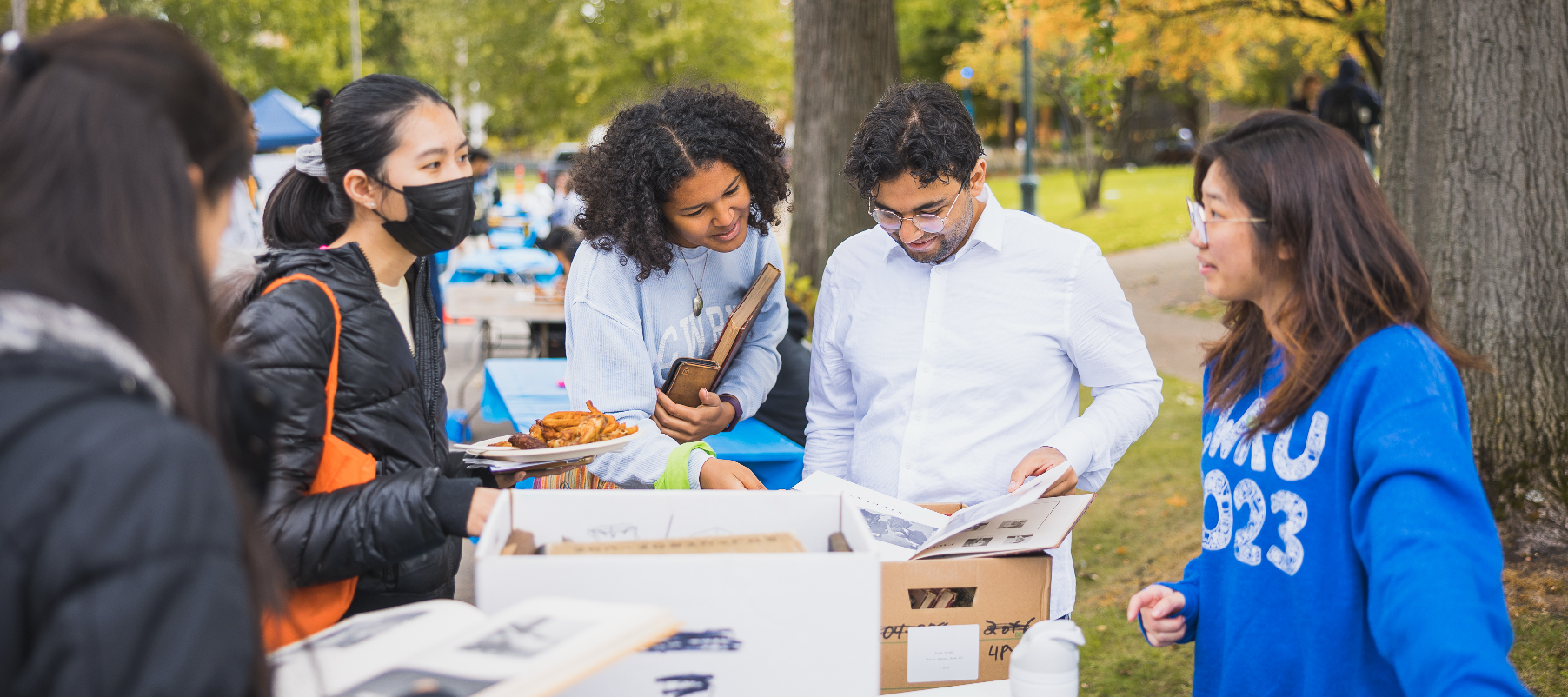 Group of diverse community members gathered outdoors at a campus event, reviewing printed materials and engaging in conversation at a table display.