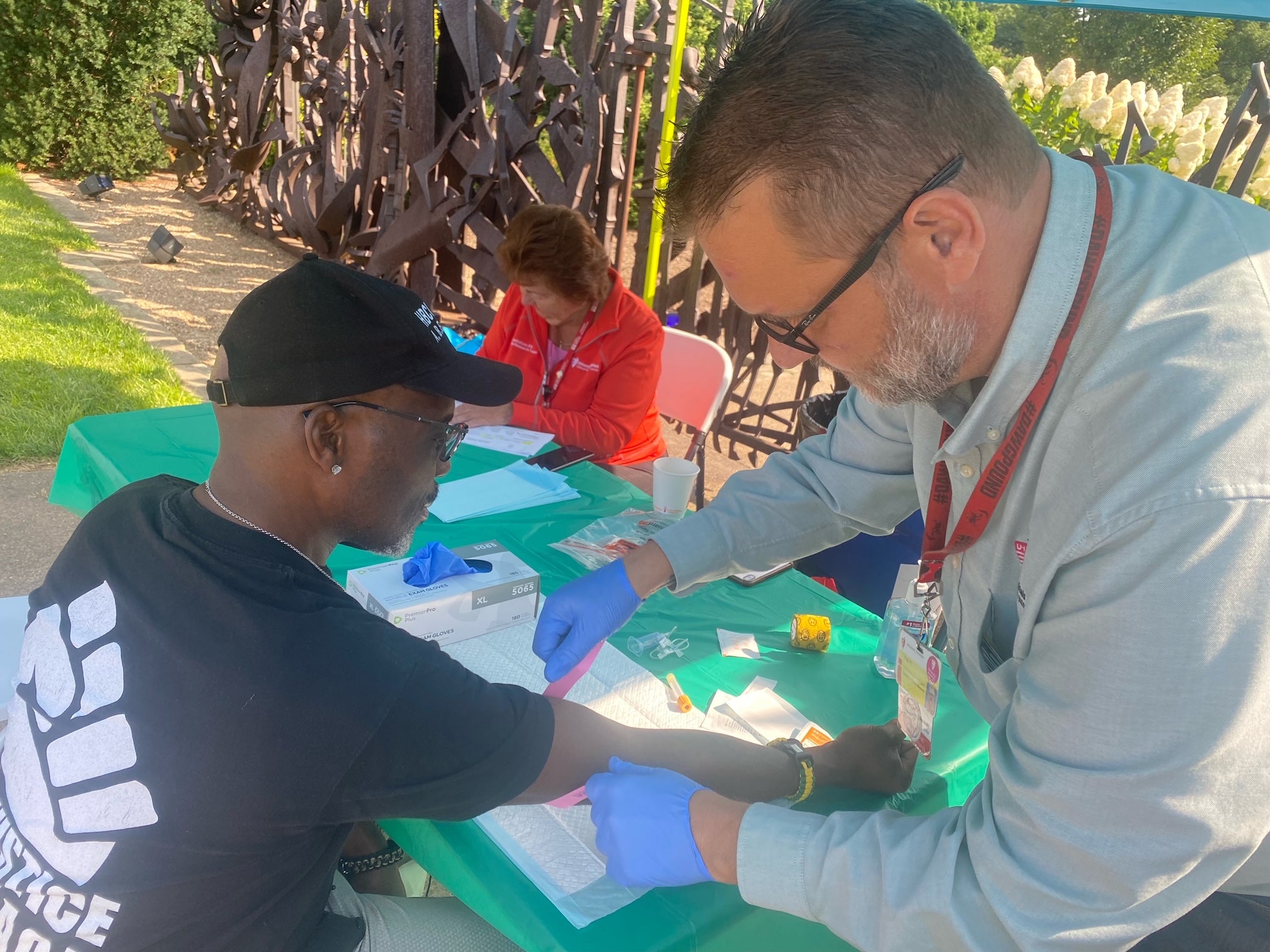A healthcare professional wearing blue gloves collects a blood sample from a community participant at an outdoor health screening event while staff assist other attendees in the background.