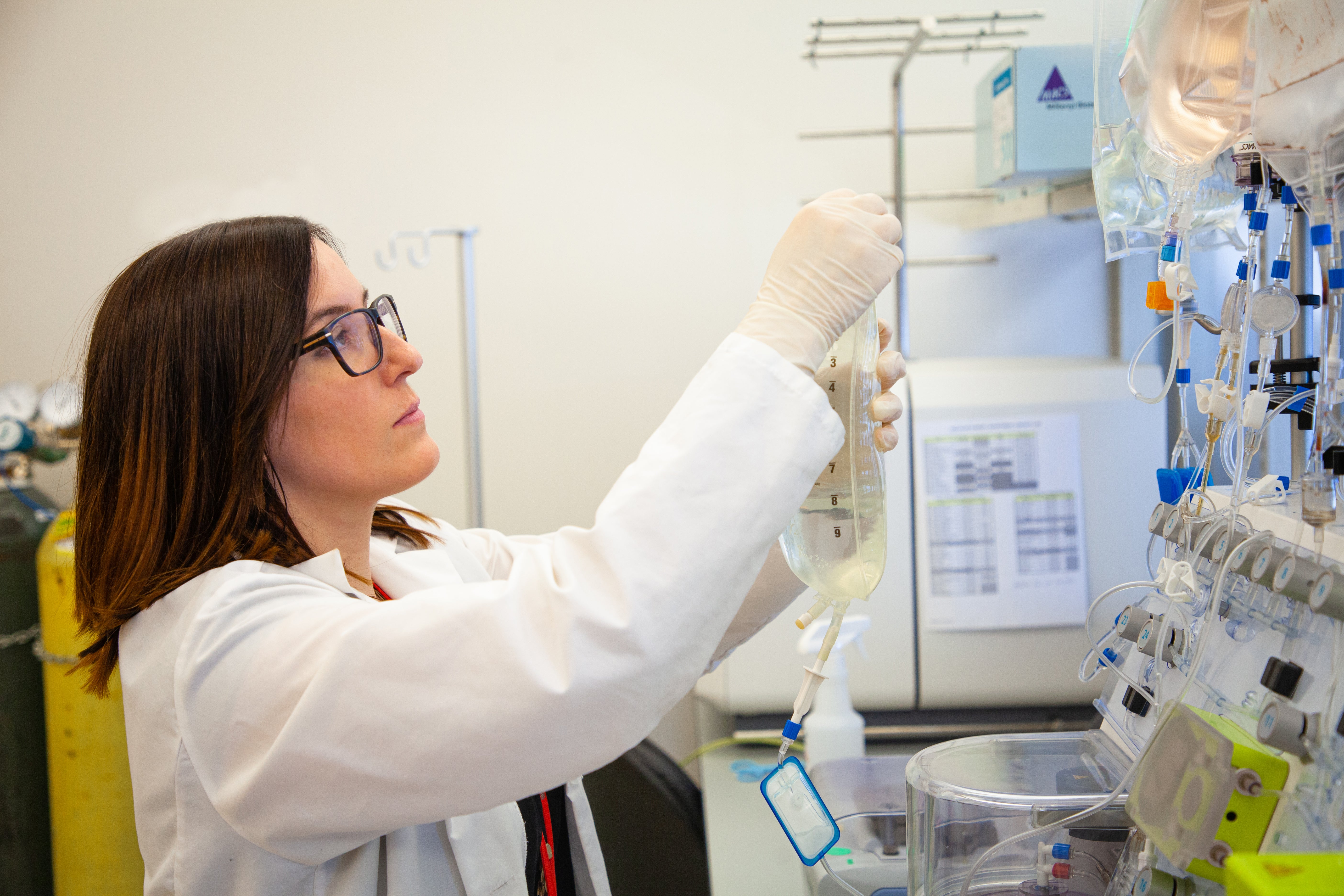 A laboratory researcher wearing a white lab coat and gloves adjusts an intravenous fluid bag connected to specialized laboratory equipment used for cell therapy or biomedical research.
