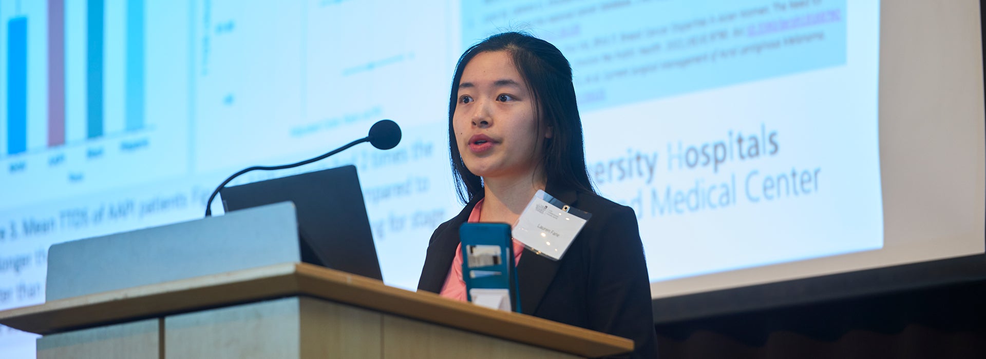Student researcher presents scientific findings at a podium during a research conference, with presentation slides displayed on a screen behind her.
