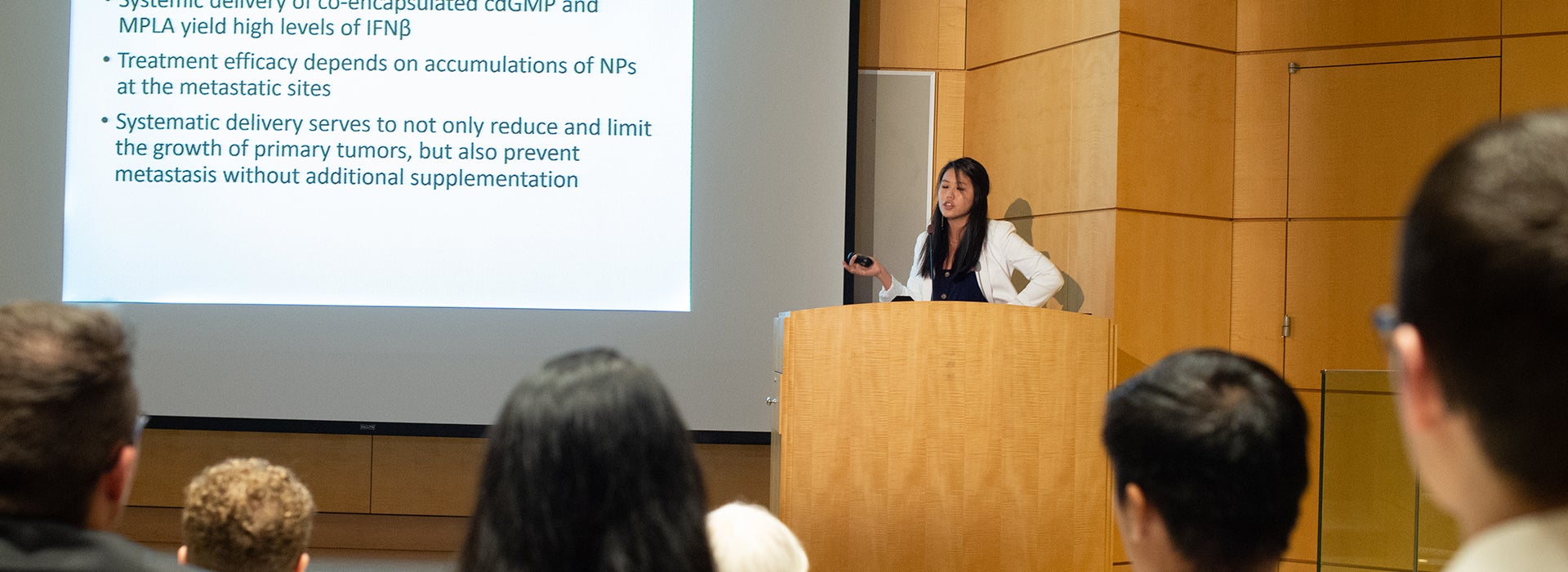 Student researcher presents scientific findings from a podium while speaking to an audience in a university lecture hall. A research presentation slide is displayed on a large screen behind the speaker.