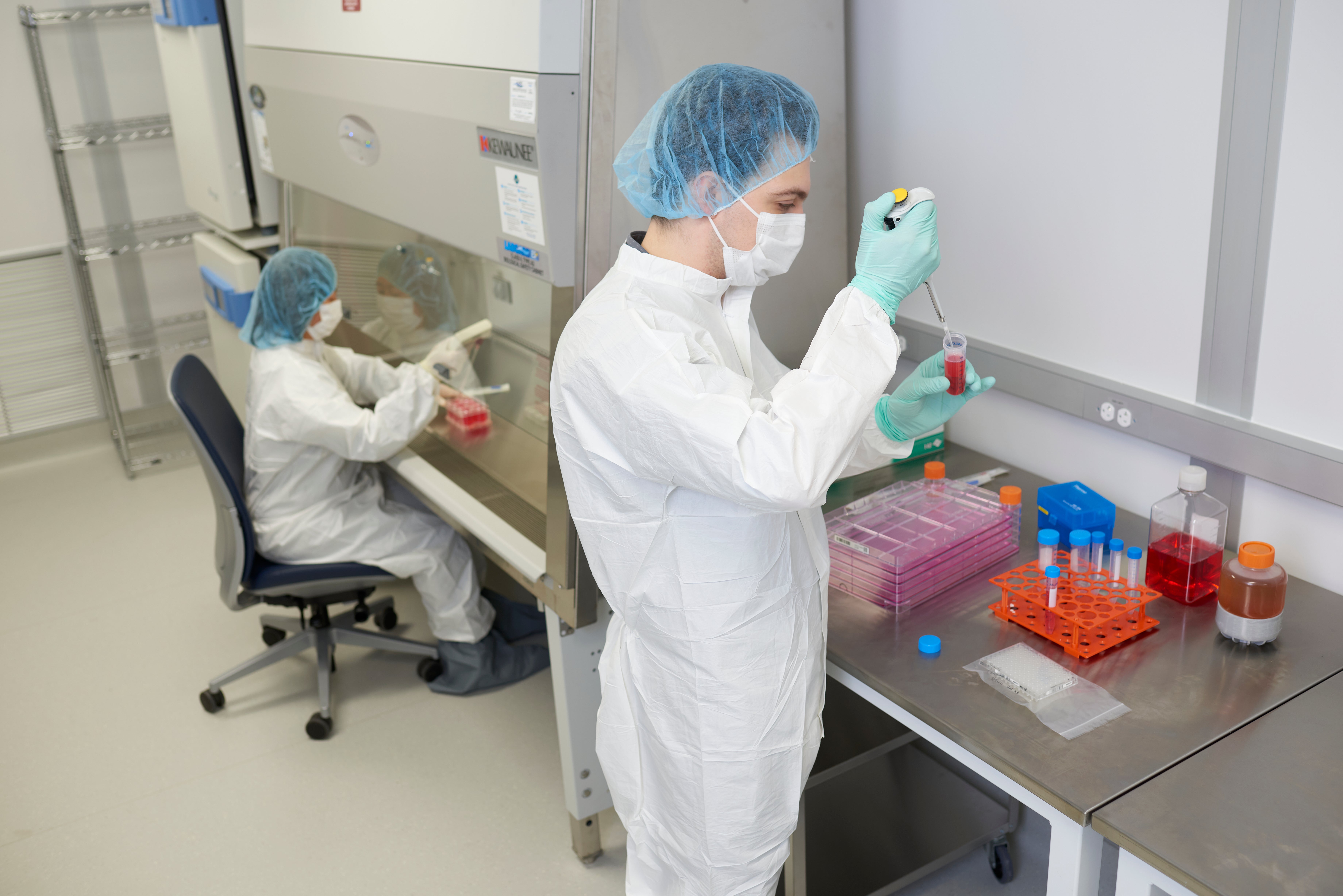 Two researchers wearing protective gowns, masks, gloves and hair covers work with red liquid samples in a laboratory.