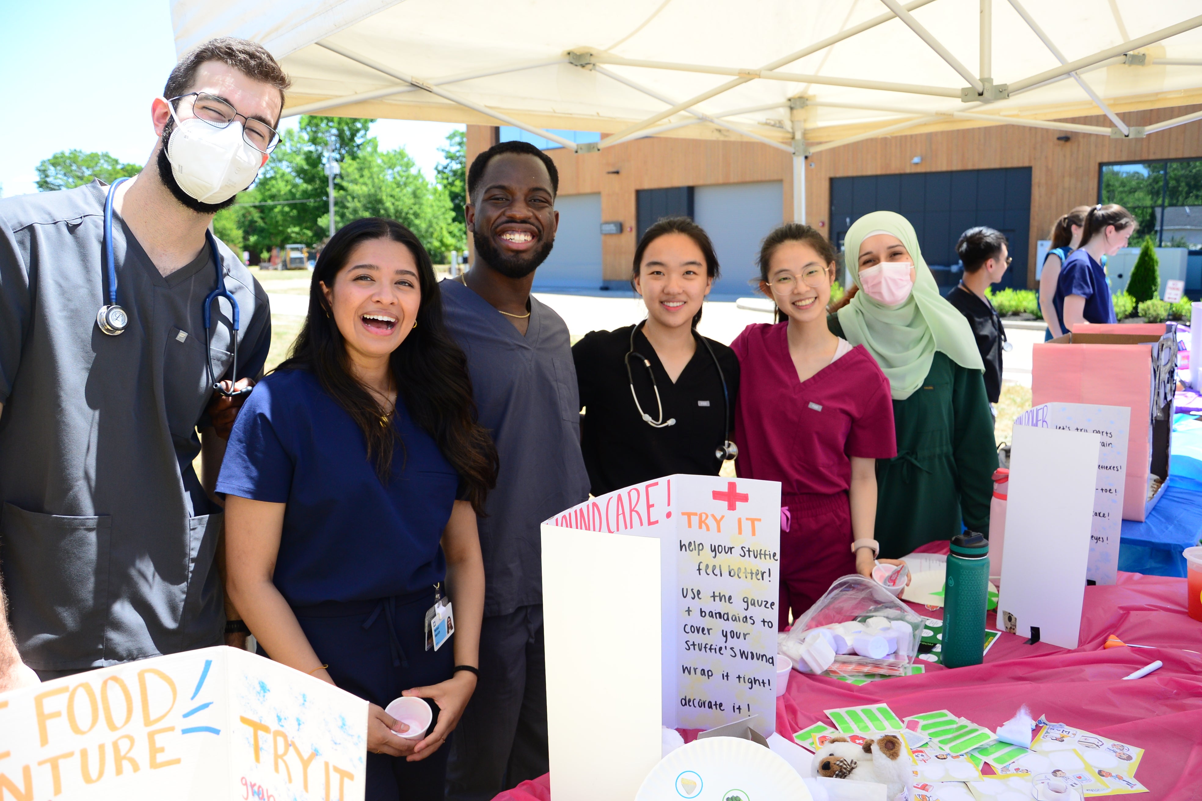 Six people wearing medical scrubs stand behind an activity table at an outdoor community event.