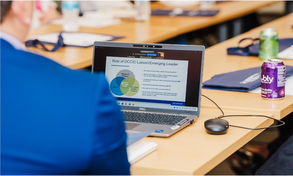 A laptop displays a presentation during a meeting, with attendees seated around a conference table.