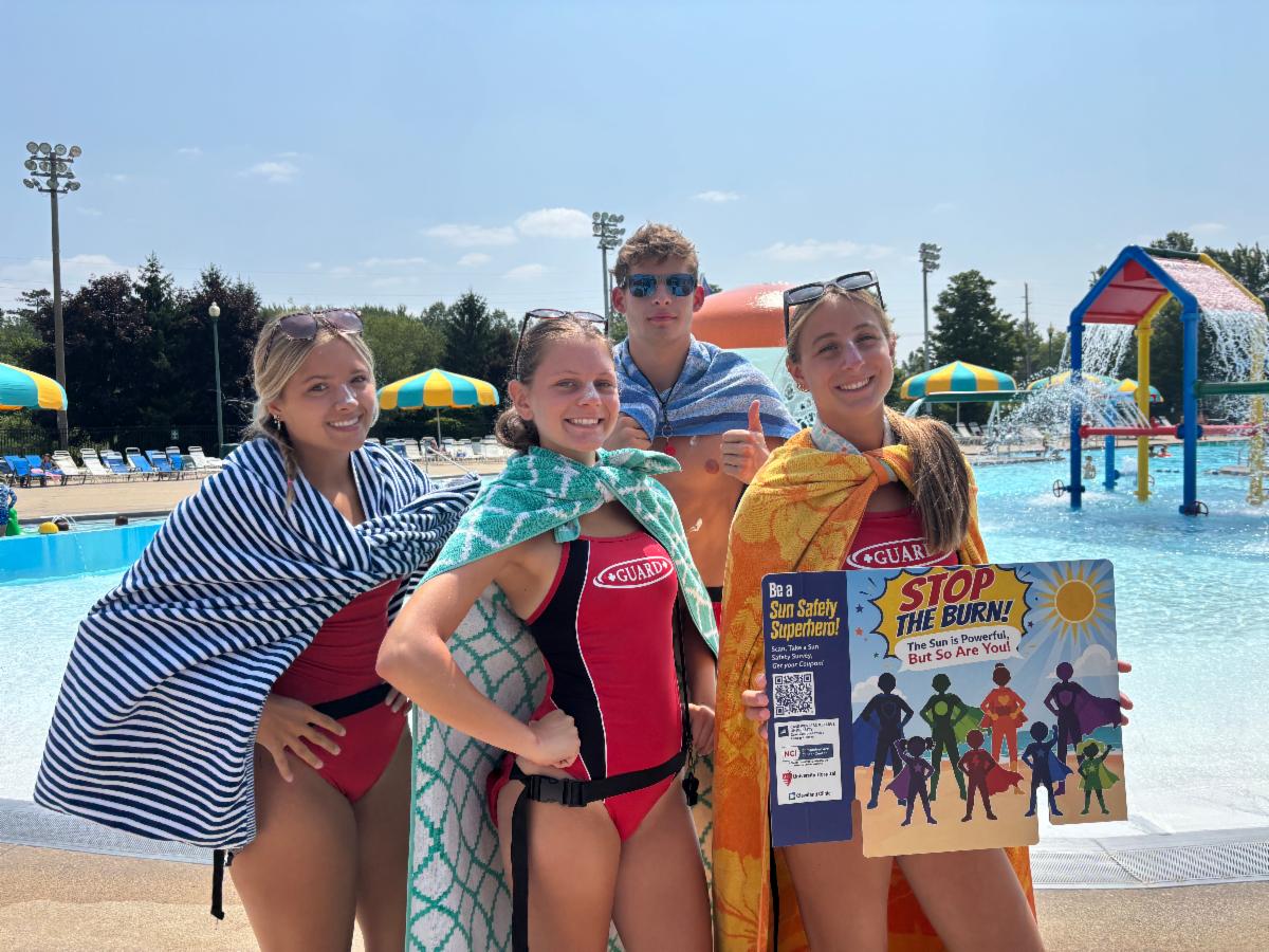 Lifeguards at an outdoor pool pose with a Stop the Burn sun safety sign promoting sunscreen use and skin cancer prevention.