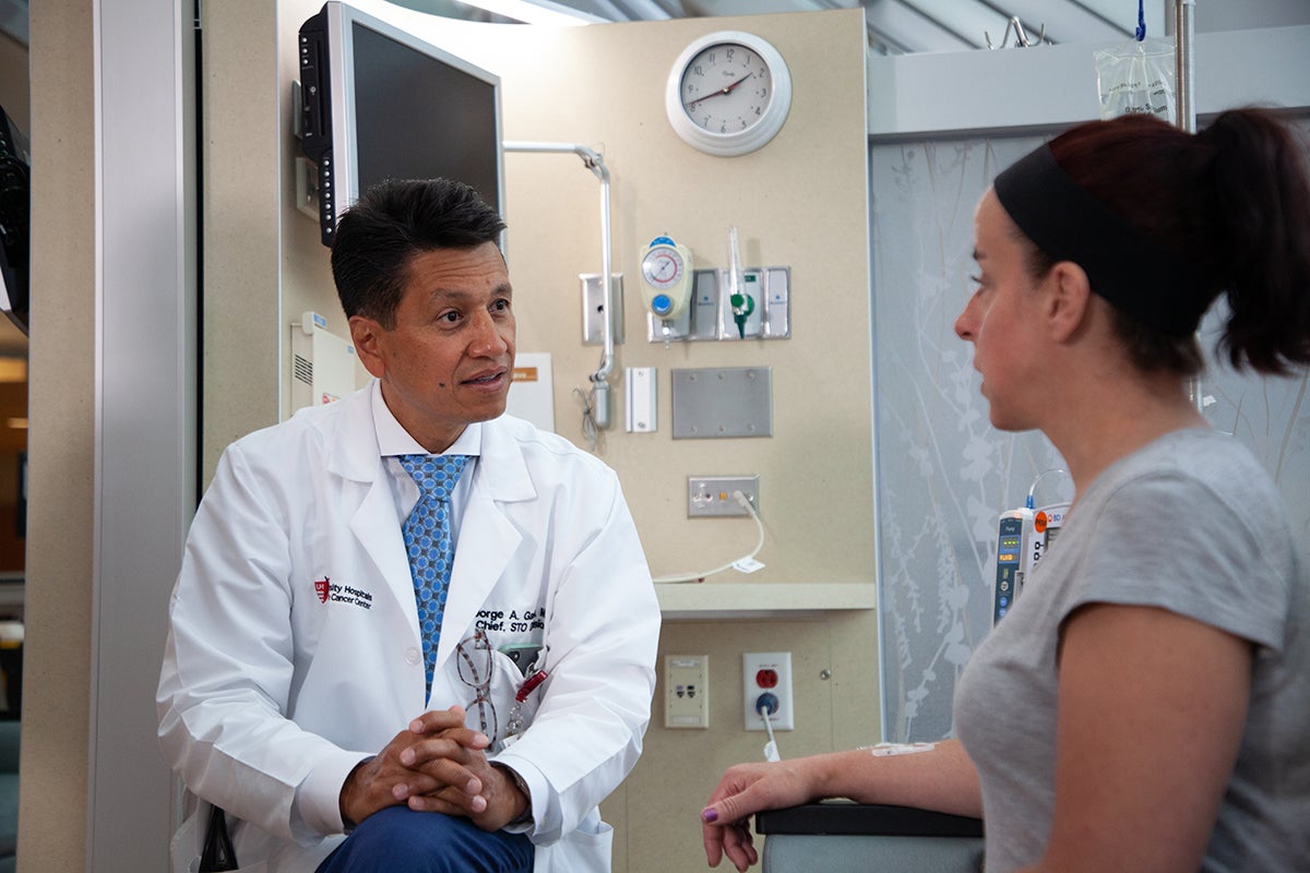 A physician in a white coat speaks with a seated patient in a clinical exam room.