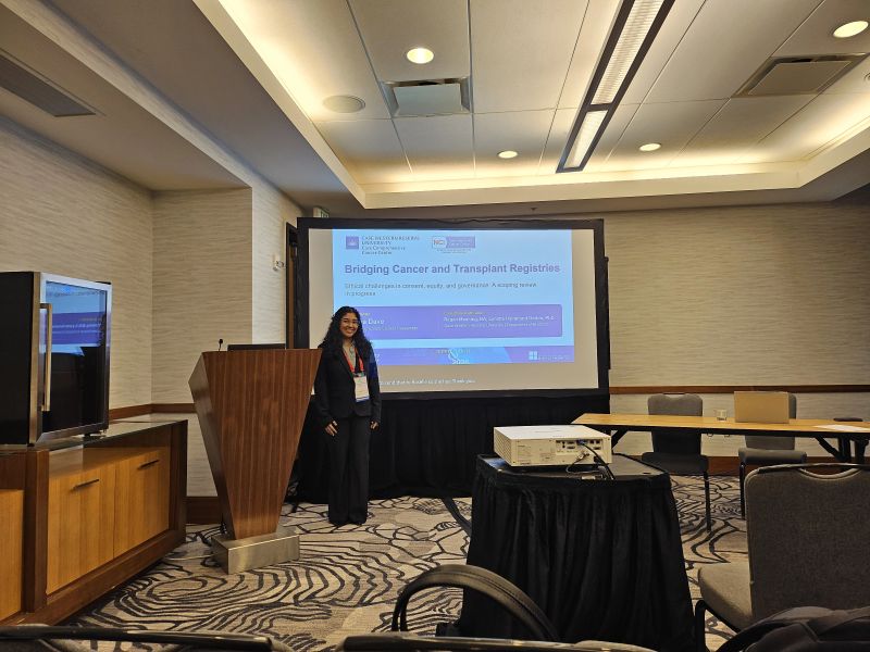 A researcher stands beside a projection screen displaying a presentation titled “Bridging Cancer and Transplant Registries” in a conference room.