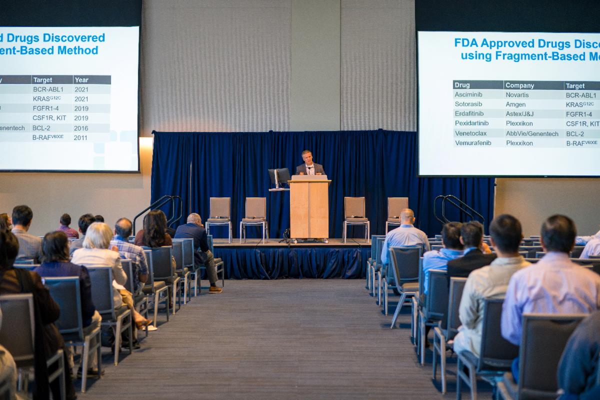 A speaker presents research on stage to attendees during the Case Comprehensive Cancer Center Annual Scientific Retreat, with presentation slides displayed on large screens.