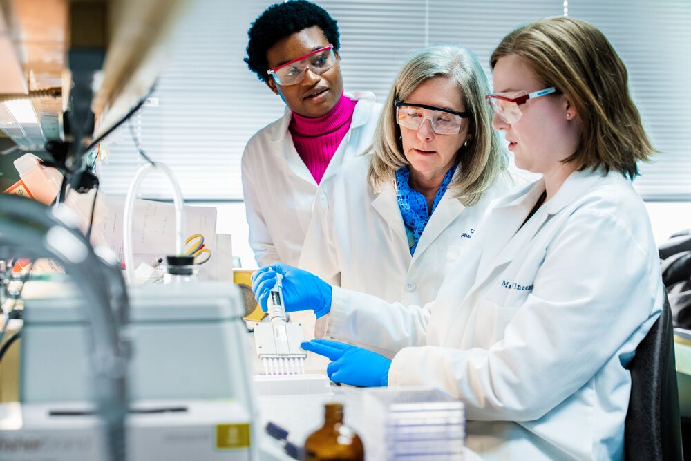Members of the Keri lab in lab coats gather around a scientific work bench with pipettes