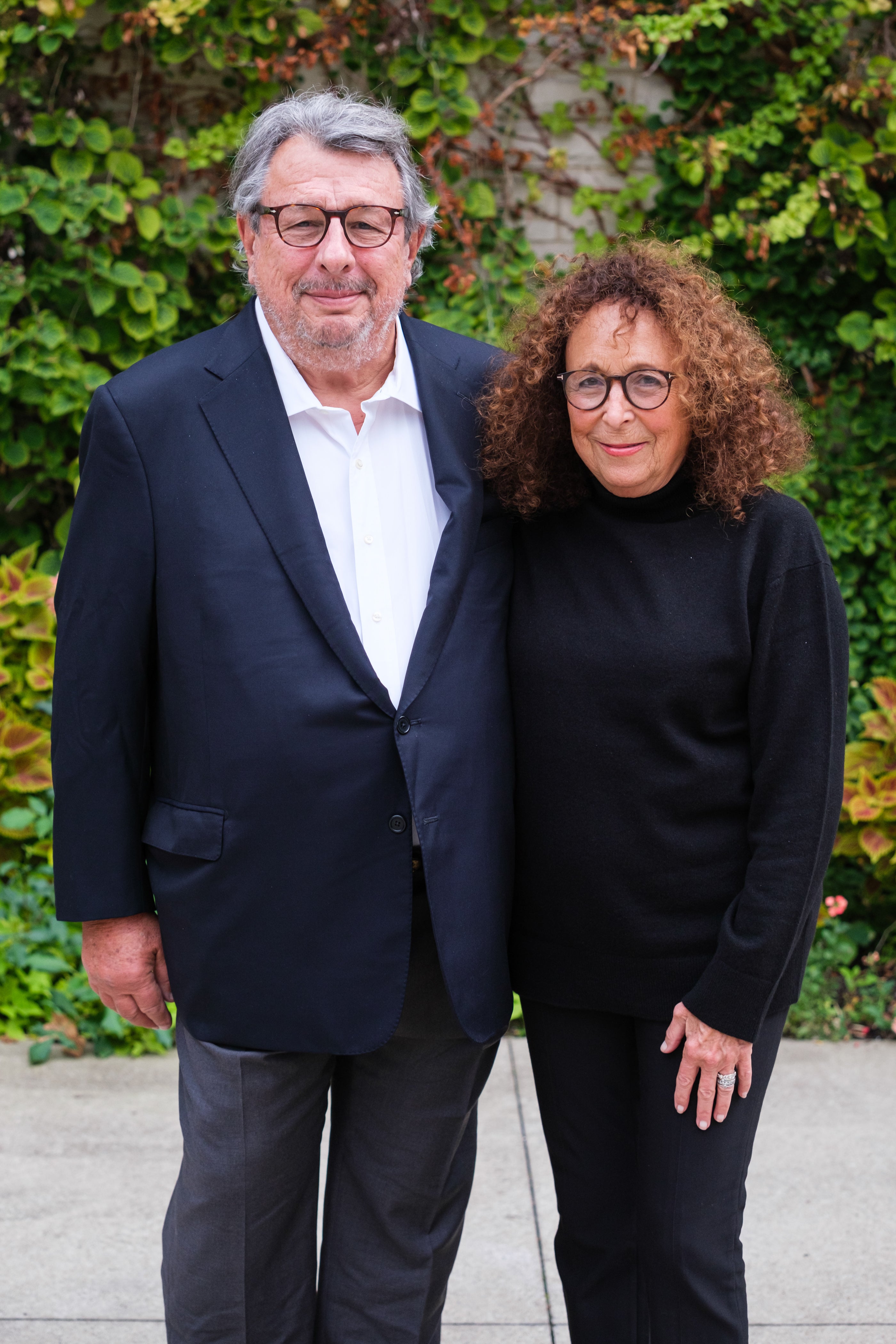 Mark and Linda Taxel stand together, smiling for a portrait outdoors in front of a leafy green backdrop.
