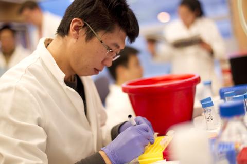 Research trainee wearing a lab coat and gloves labels laboratory samples while working at a laboratory bench surrounded by scientific equipment.