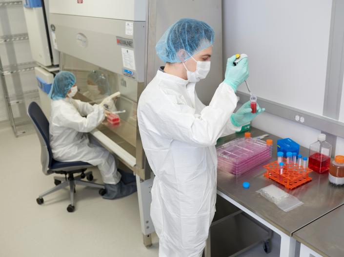 Two researchers wearing protective gowns, masks, gloves and hair covers work with red liquid samples in a laboratory.
