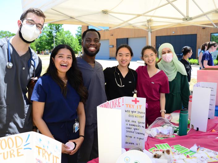 Six people wearing medical scrubs stand behind an activity table at an outdoor community event.