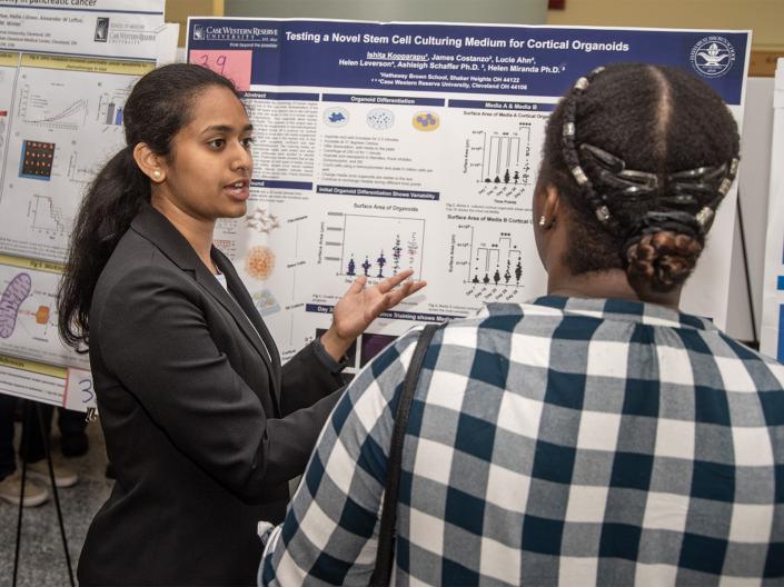 A researcher presents a scientific poster on stem cell culturing for cortical organoids to an attendee during a poster session. The presenter gestures toward charts and diagrams on the poster while explaining the research, with additional research posters displayed in the background.