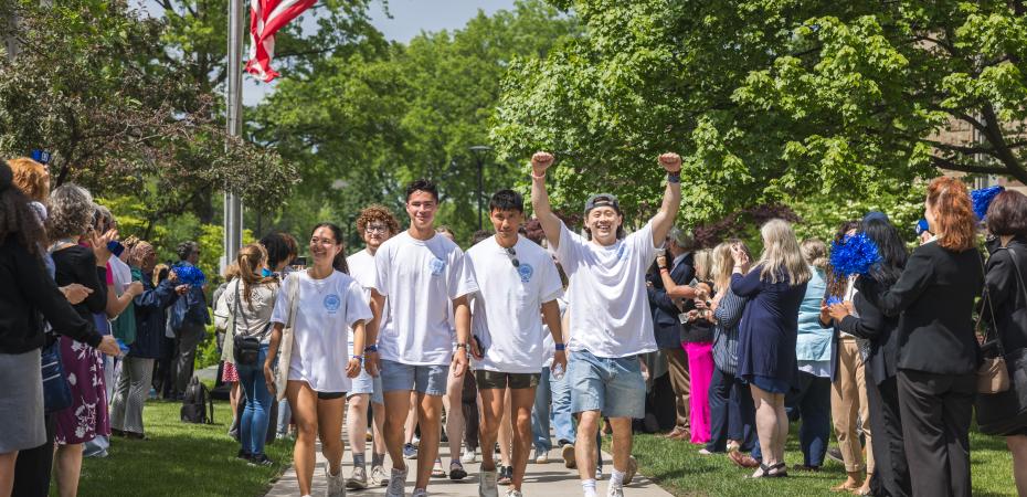 Photo of students walking down a cement pathway celebrating with arms up as staff and faculty clap / congratulate them in their graduation achievements