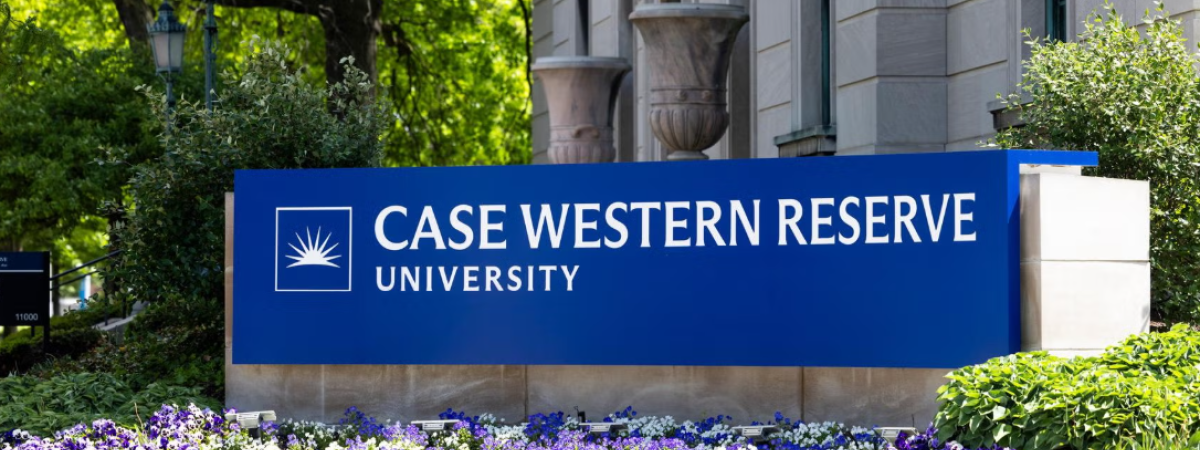A blue sign that says Case Western Reserve University sits in front of a stone building with purple flowers in front.