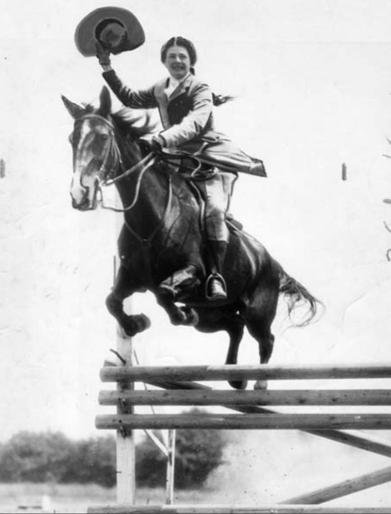 Adele Parker, astride a horse and waving her hat jumps a three-rail barrier.