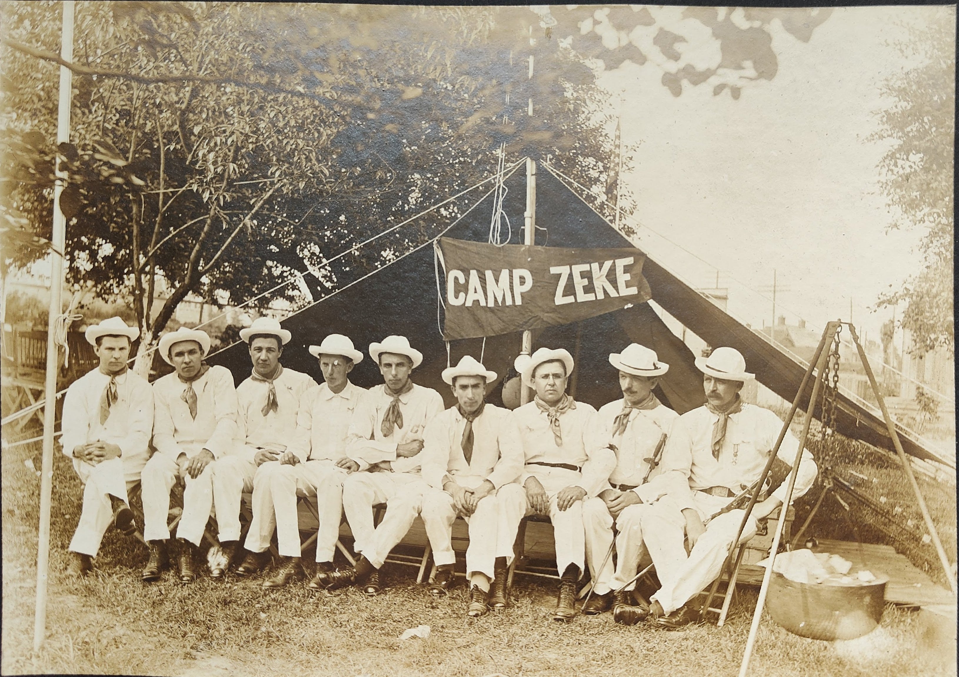 Nine men dressed in white uniforms with white hats sit on a bench in front of tent at Camp Zeke in Cleveland
