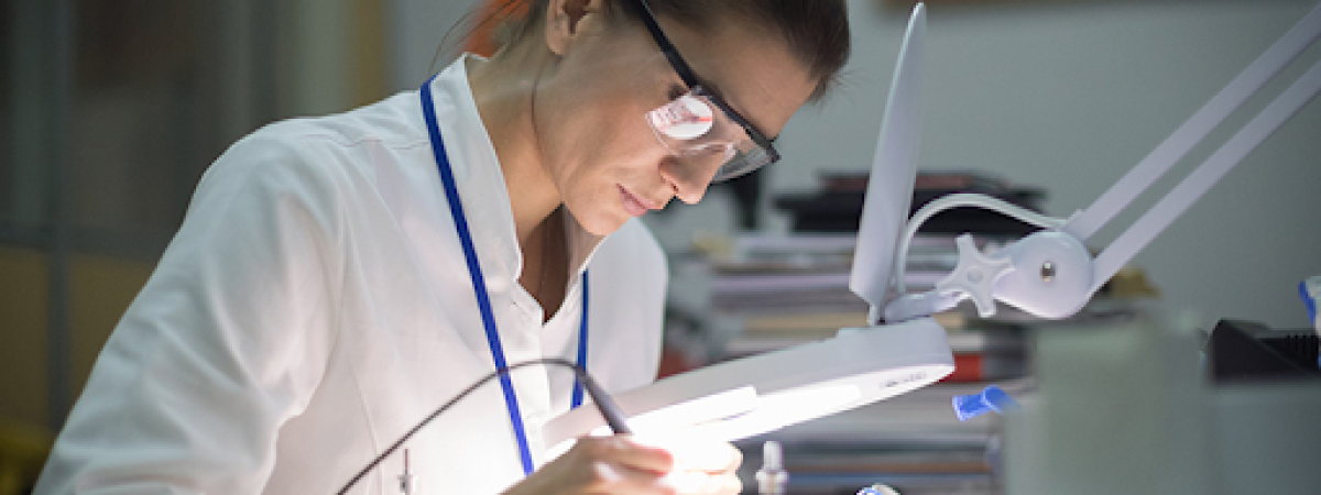 A woman in a lab coat and safety glasses solders an electronic circuit board under a magnifying lamp.
