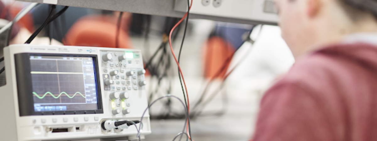 A student works at a lab bench with an oscilloscope displaying a wavy green line.