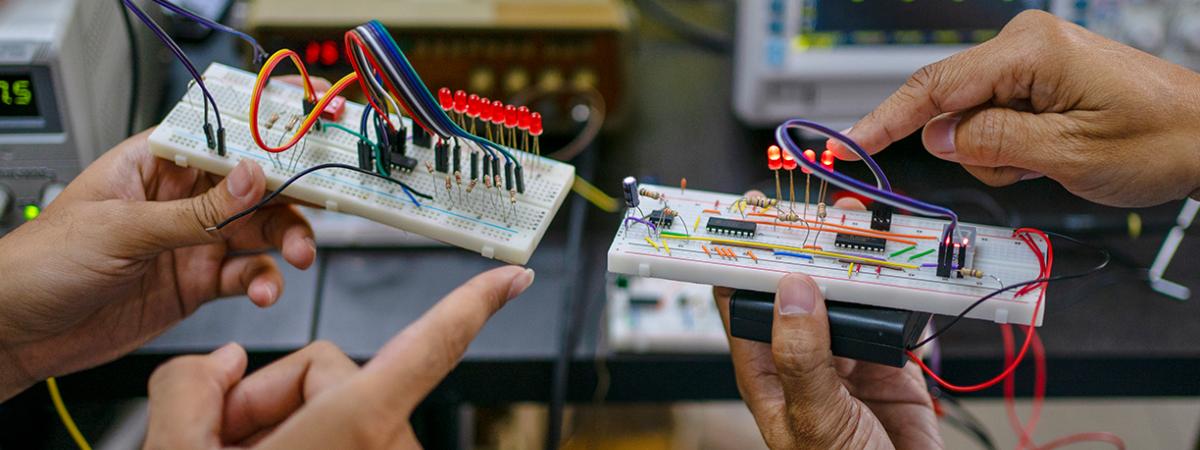 A close-up of two people holding and pointing at electronic circuit breadboards with wires