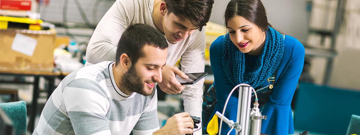 Three engineering students are gathered around a table, engaged in a discussion while assembling a small robotic vehicle.