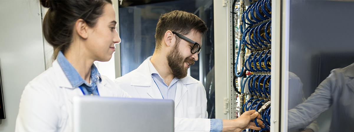 Two engineers in lab coats work together in a server room, with one holding a laptop.