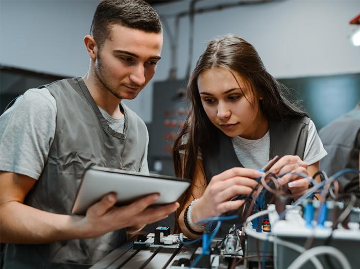 Two focused engineering students, a male and a female, working together on a complex electronic project with various wires, with the male student holding a tablet.