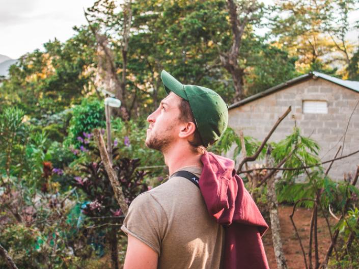 A college student wearing a green baseball hat looks at the sky while standing in a garden near a brick building