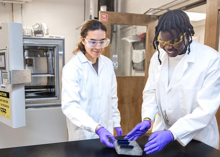 two students in a lab with purple gloves with one student wearing goggles