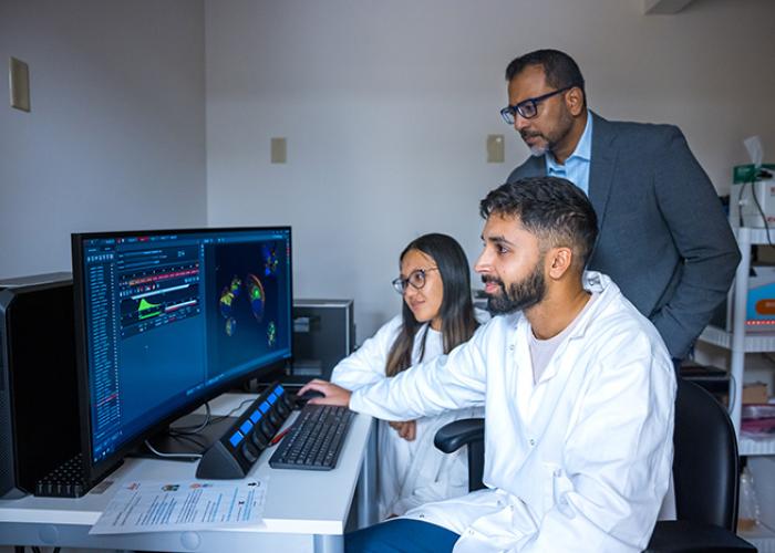 One person standing and two people sitting in front of a computer