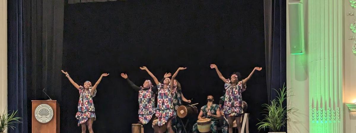 Picture of four dancers dancing on stage with drummers behind them. 