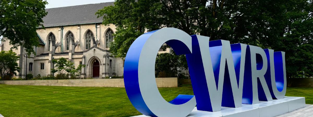 A large 3D sign spelling out "CWRU" in silver and blue stands on a campus lawn in front of a historic, Gothic-style stone building.