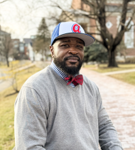 Headshot for Dr. Bennett, he is wearing a grey sweater, and a bow tie and hat. 