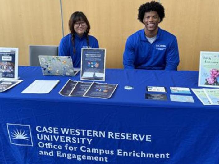 Two people sitting behind a blue-clothed information table for Case Western Reserve University's Office for Campus Enrichment and Engagement, smiling at the camera with flyers and brochures.
