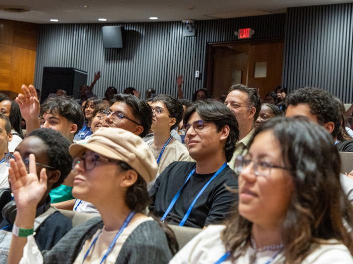 An audience of diverse students sitting in a lecture hall or auditorium, with several individuals raising their hands to participate or ask questions during a presentation.