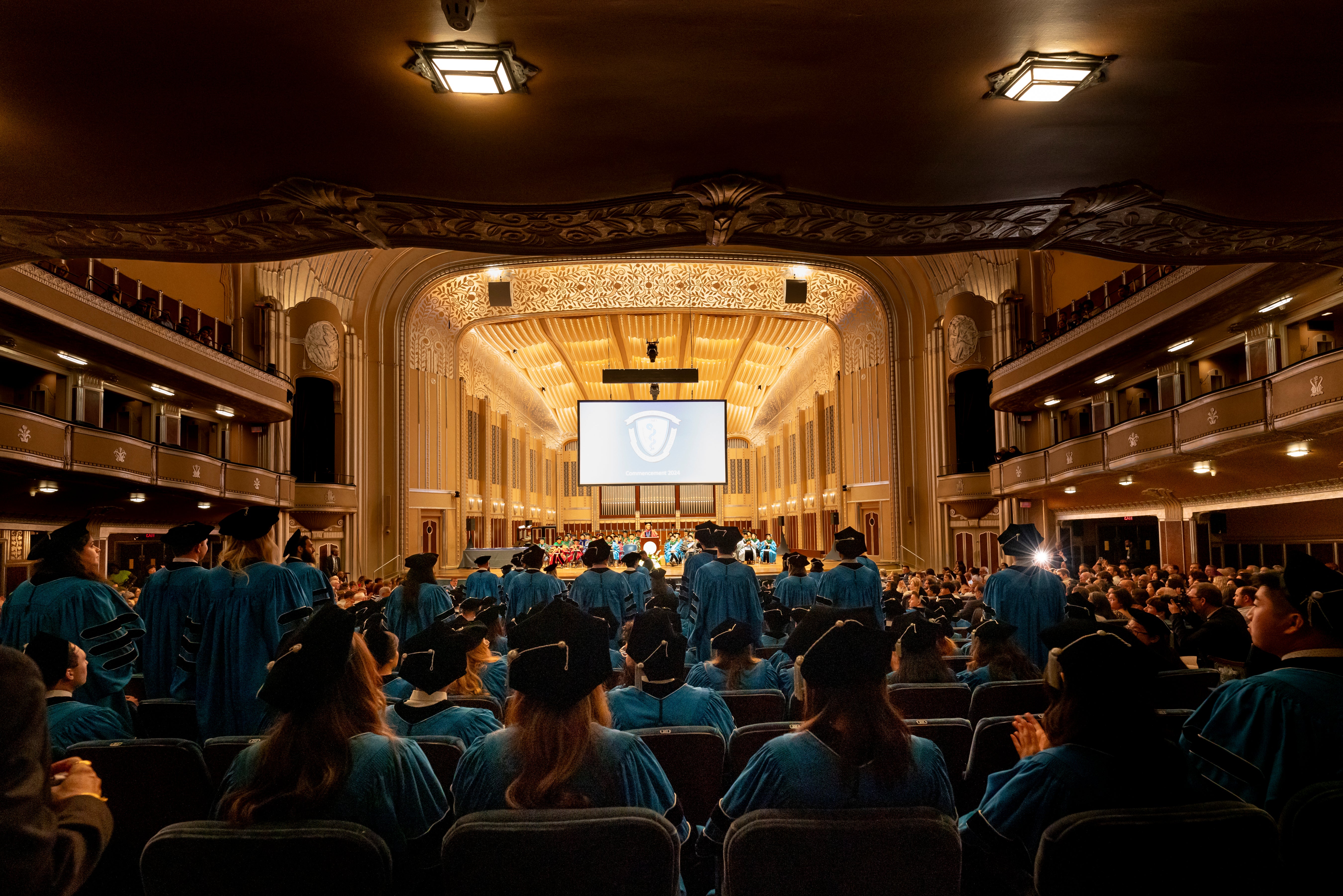 Students sitting in a bog auditorium with their graduation caps and gowns on. 