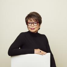 Lady with short hair, glasses, and black turtleneck standing behind a white podium.