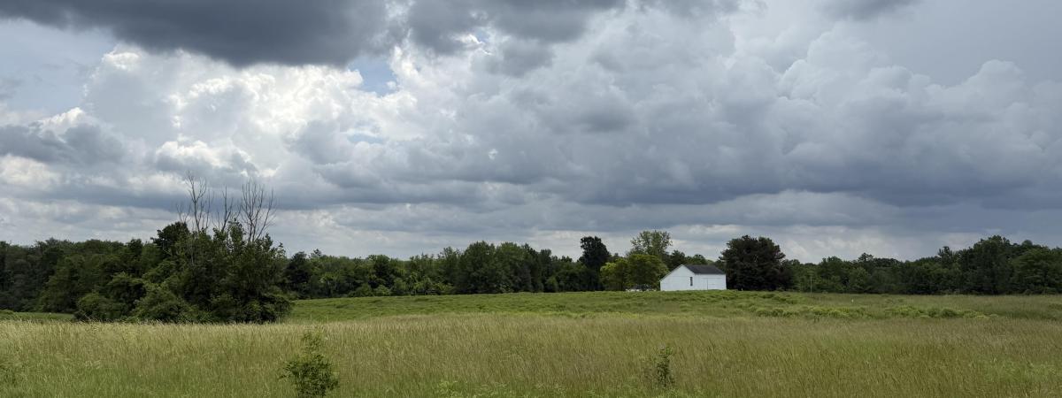 A stormy sky hangs over a green field with a white barn in distance