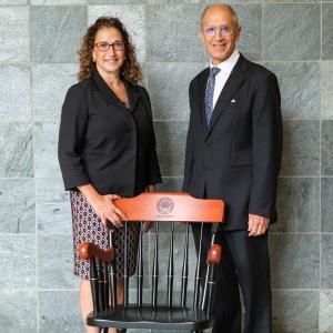 Lia Logio and Stan Gerson stand behind a CWRU-branded chair while wearing formal attire and smiling at the camera in front of a gray marble tile wall