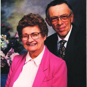 Ruth Barber and David Moon smile at camera in front of a plain portrait backdrop and a floral arrangement while wearing formal attire.