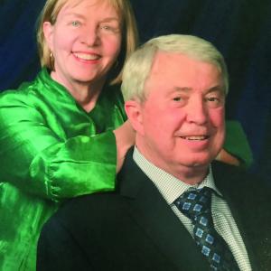 Sally Bonacker Warburton and Phillip Warburton smile at camera in front of a plain, dark portrait backdrop while wearing formal attire