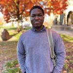 Chibunnam Gerald Onyedika stands in the Case Quad in fall, wearing a collared shirt and gray sweater with a backpack on one shoulder